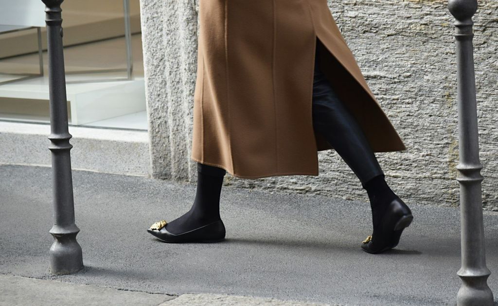 Black flat shoes worn with black tights and a camel coat on a city pavement.